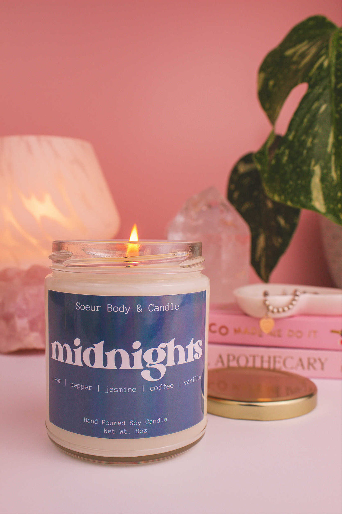 Midnights candle lit, with a stack of books and a jewelry dish in the background, set against a lamp, plant, and crystals; fragrance notes: pear, pepper, jasmine, coffee, and vanilla.