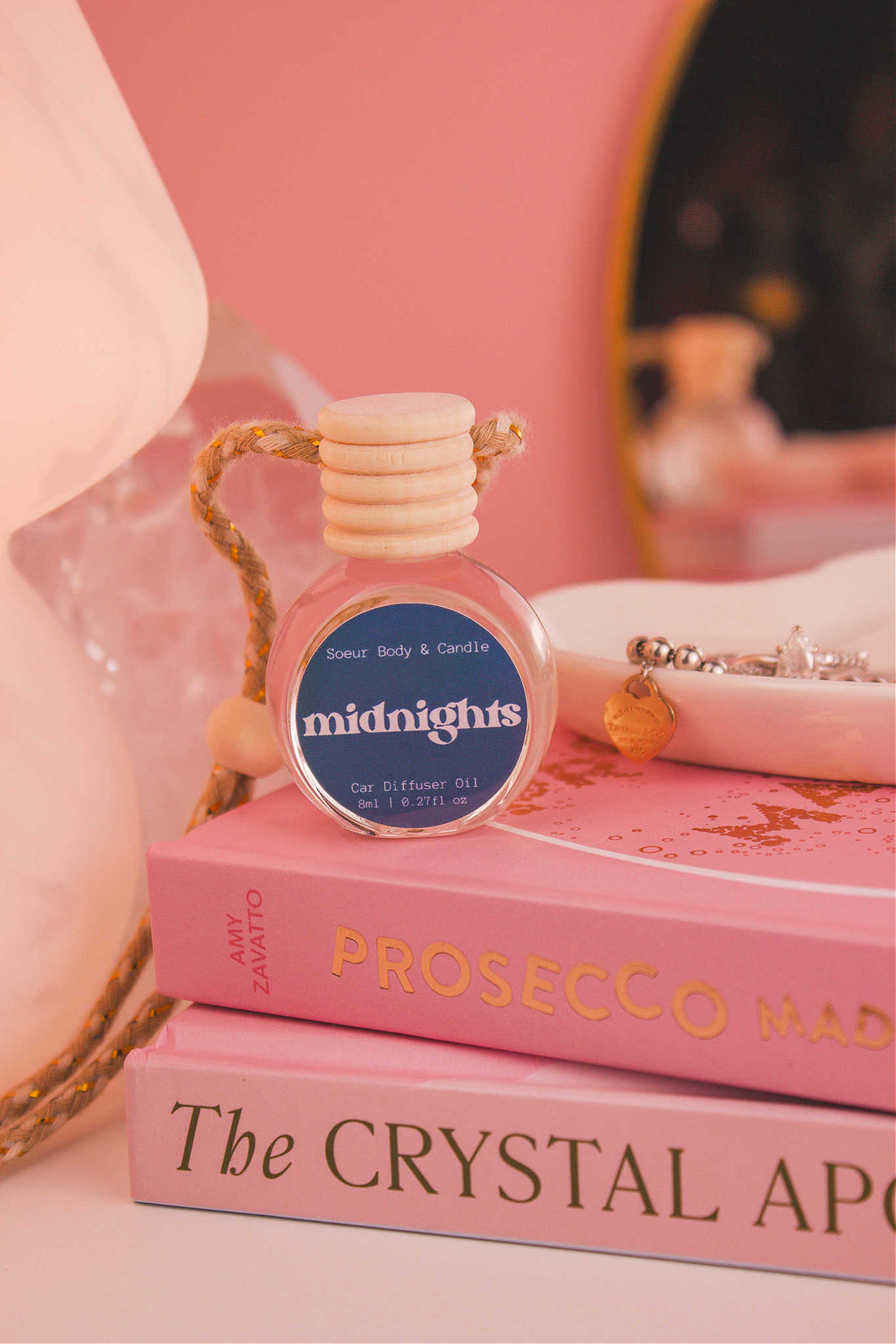 Jaded diffuser sitting on top of a stack of books with a jewelry dish in the background, set against a lamp, mirror, and crystals; fragrance notes: pear, pepper, jasmine, coffee, and vanilla notes.