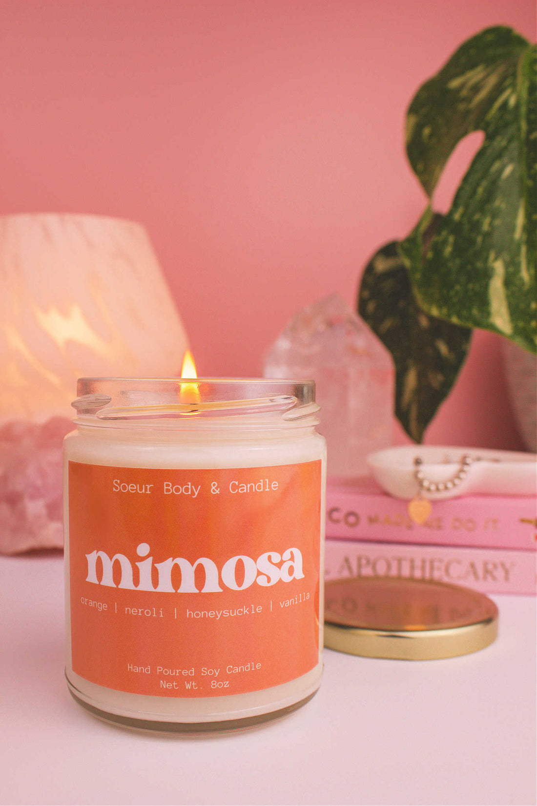 Close-up of Mimosa candle lit, with a stack of books and a jewelry dish in the background, set against a lamp, plant, and crystals; fragrance notes: orange, neroli, honeysuckle, and vanilla.