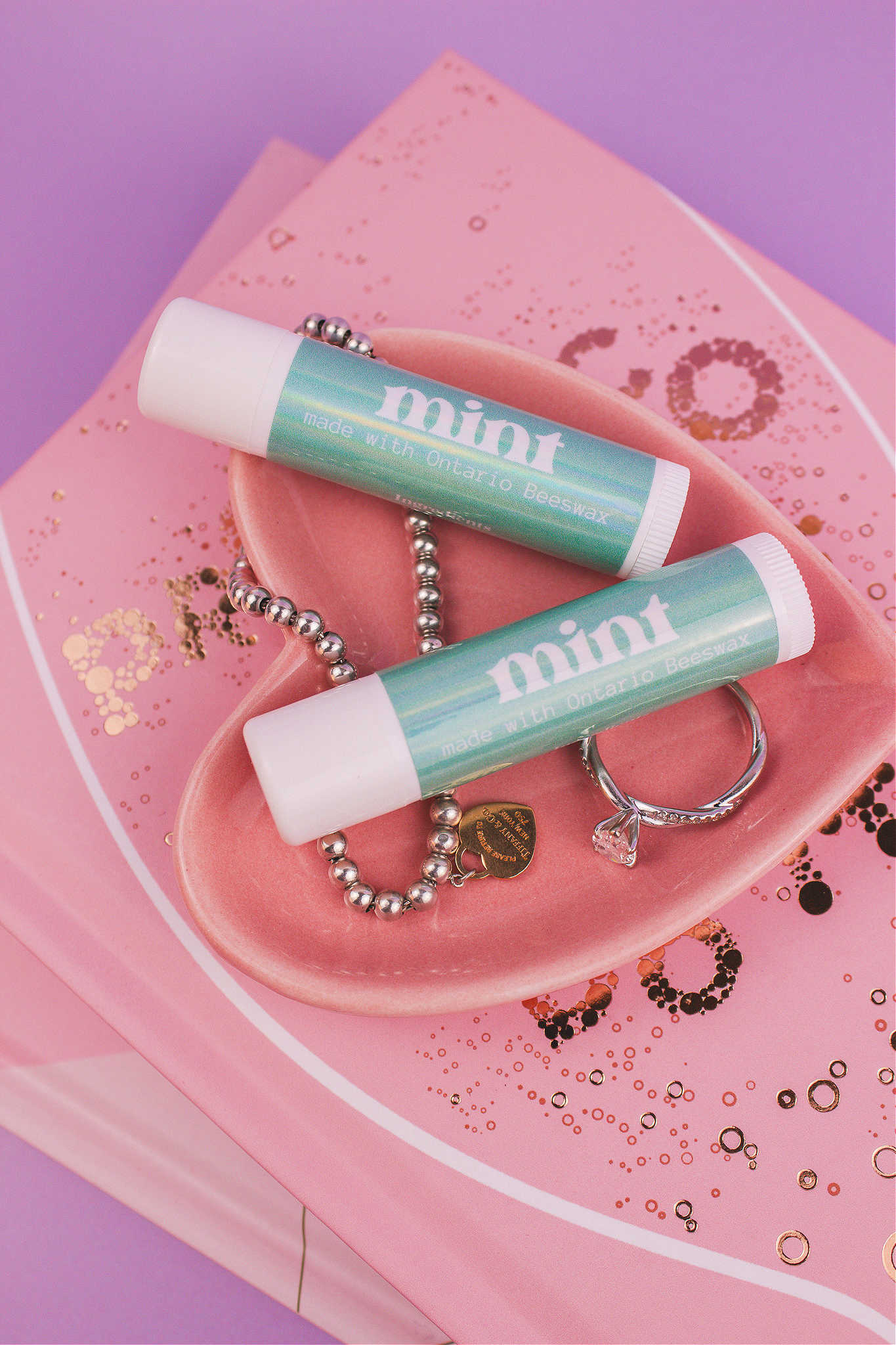 Two Mint flavoured lip balms displayed in a jewelry dish with a ring and bracelet, sitting on top of books.