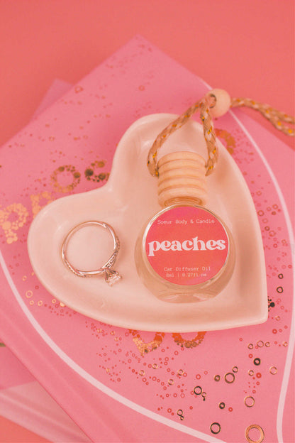 Peaches car diffuser in a jewelry dish with a ring beside it, sitting on top of books, featuring notes of orange, persimmon, jasmine, peach, and musk.