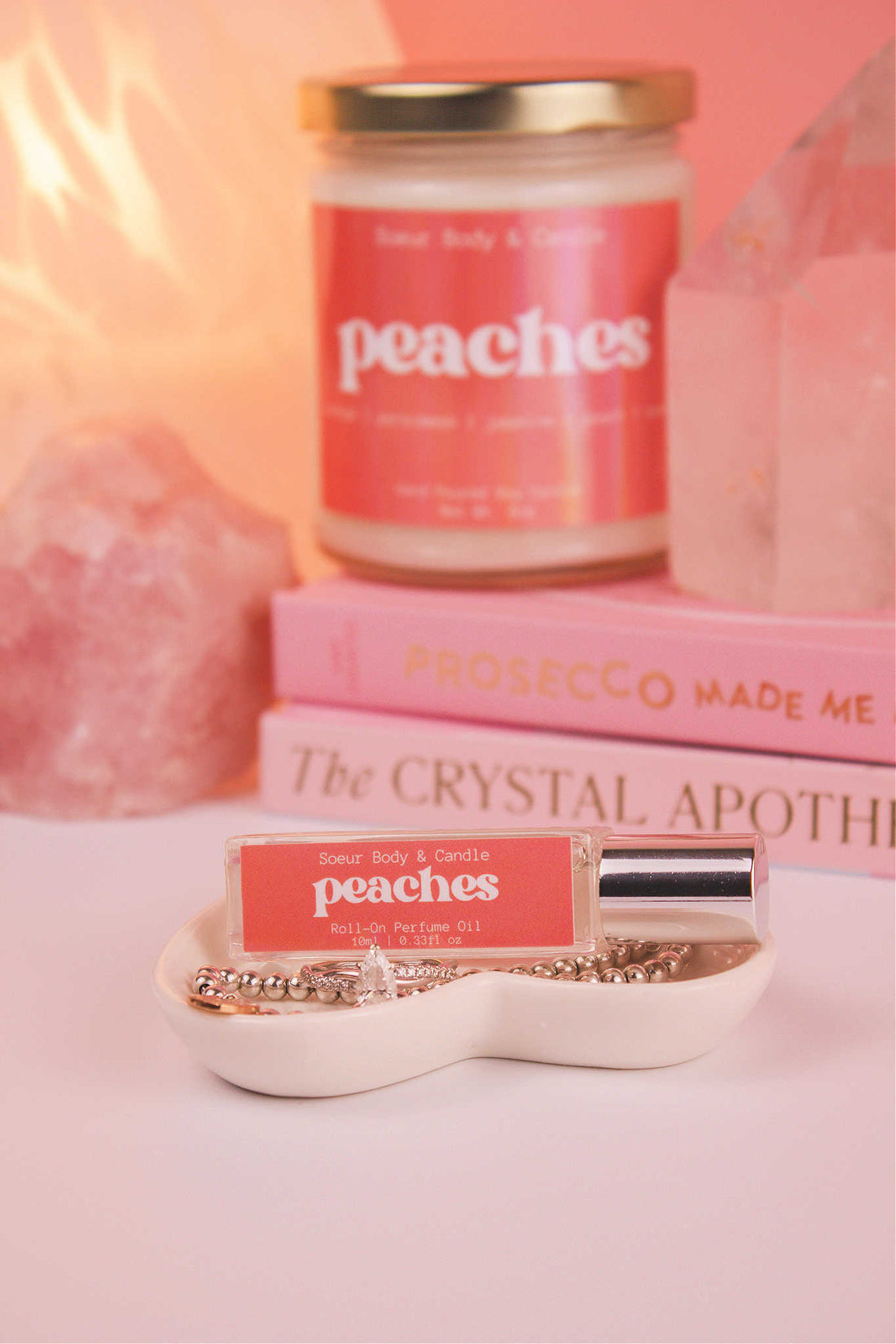 Peaches roll-on perfume elegantly set in a jewelry dish with jewelry, and coordinating candle, along with books, a lamp, and crystals in the background. Featuring notes of orange, persimmon, jasmine, peach, and musk.