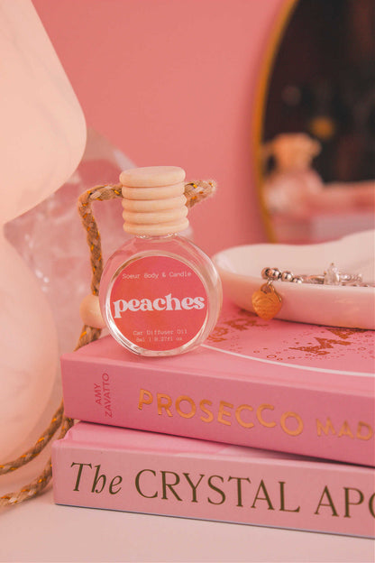 Peaches diffuser sitting on top of a stack of books with a jewelry dish in the background, set against a lamp, mirror, and crystals; fragrance notes: f orange, persimmon, jasmine, peach, and musk.