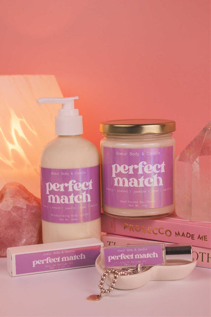 Perfect Match roll-on perfume, lotion, and candle together, along with books, a lamp, and crystals in the background, adding a cozy vibe. Featuring bold notes of cherry, almond, jasmine, rose, and vanilla.