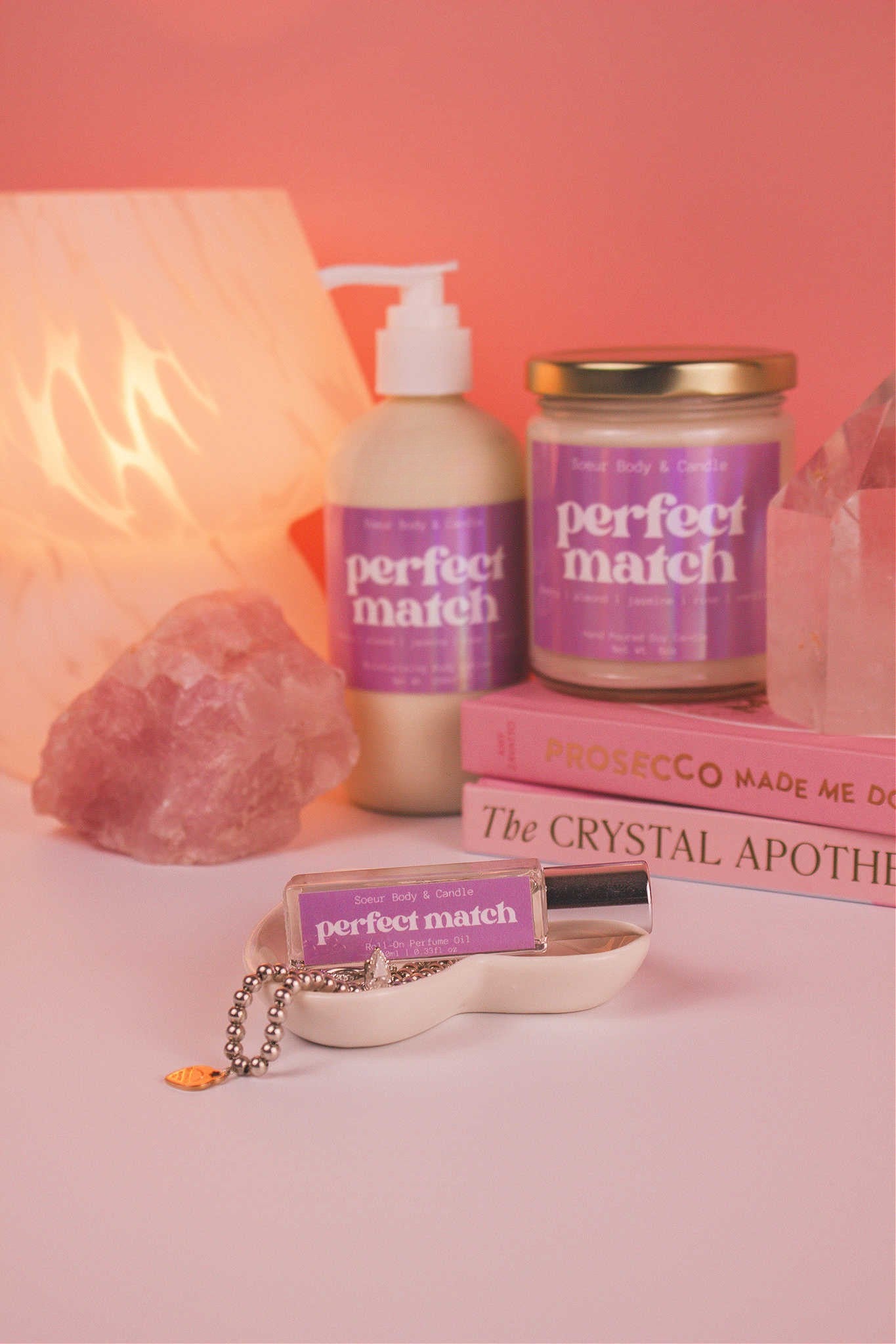 Focused shot of Perfect Match roll-on perfume in a jewelry dish with jewelry, and matching candle and lotion, along with books, a lamp, and crystals in the background. Featuring notes of cherry, almond, jasmine, rose, and vanilla.