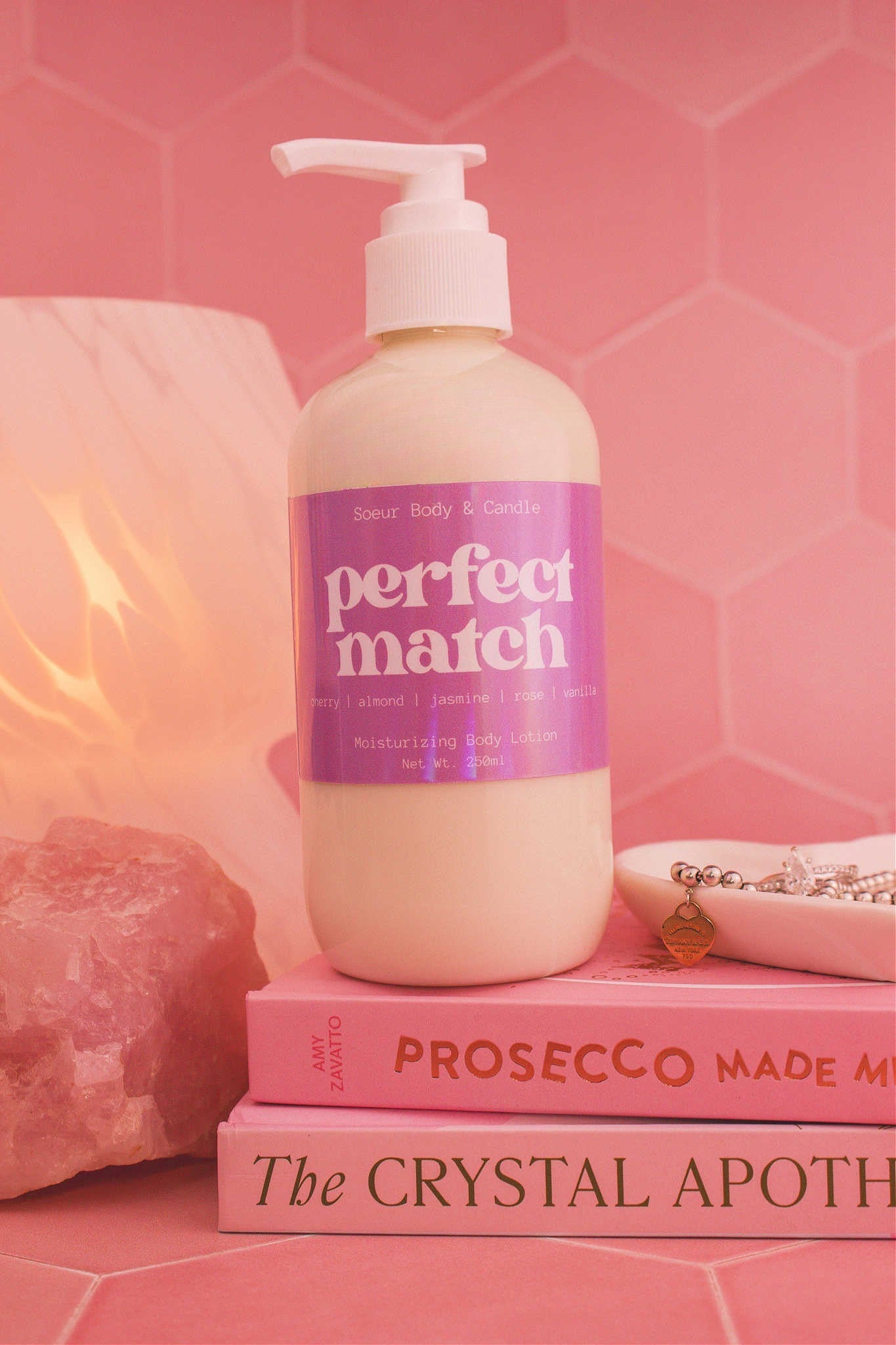 Close-up of the Perfect Match body lotion on a stack of books with a jewelry dish, lamp, and crystal in the background; fragrance notes: cherry, almond, jasmine, rose, and vanilla.