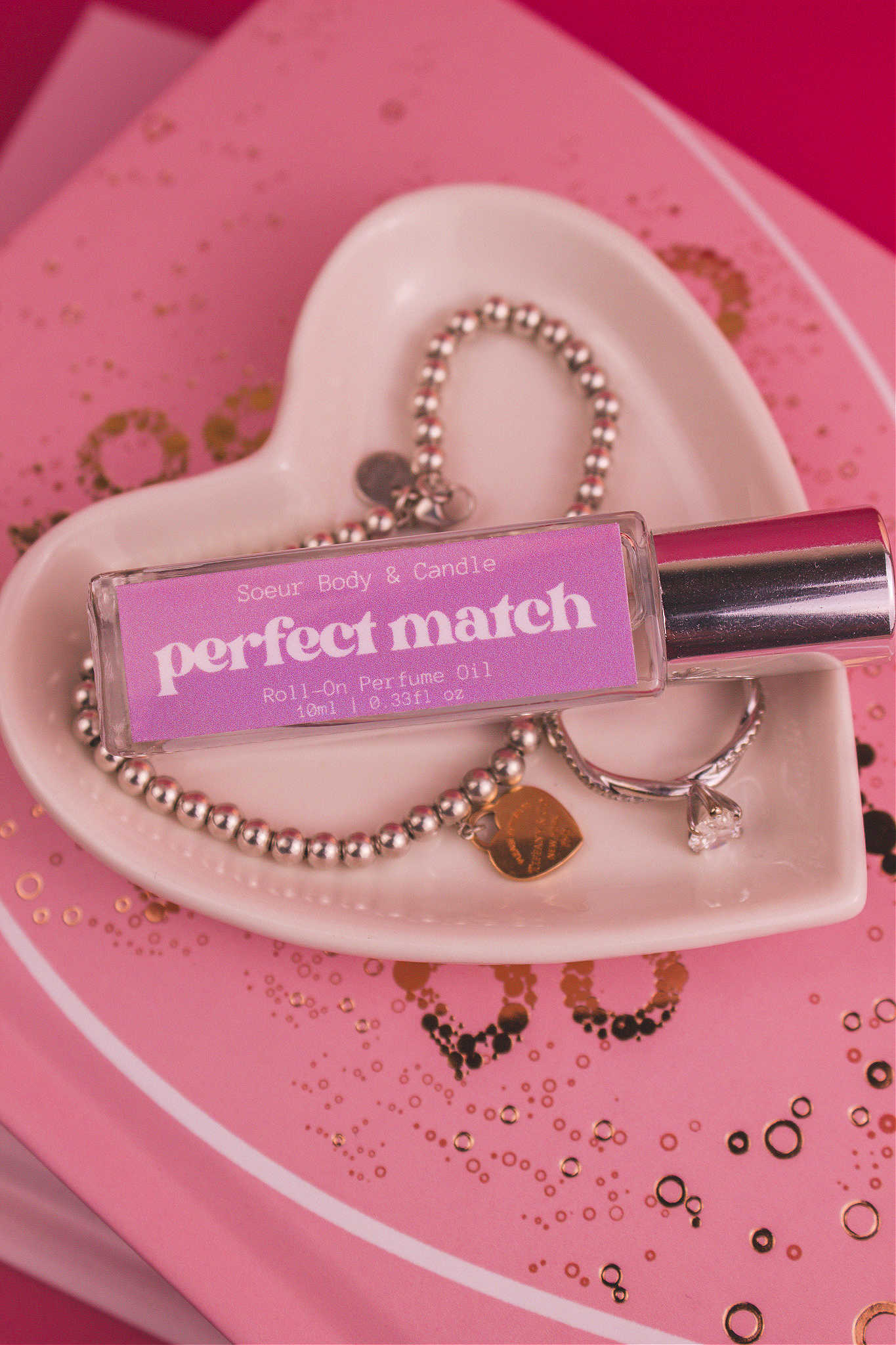 Close-up shot of Perfect Match roll-on perfume displayed in a jewelry dish beside a ring and bracelet, highlighting its blend of cherry, almond, jasmine, rose, and vanilla notes.
