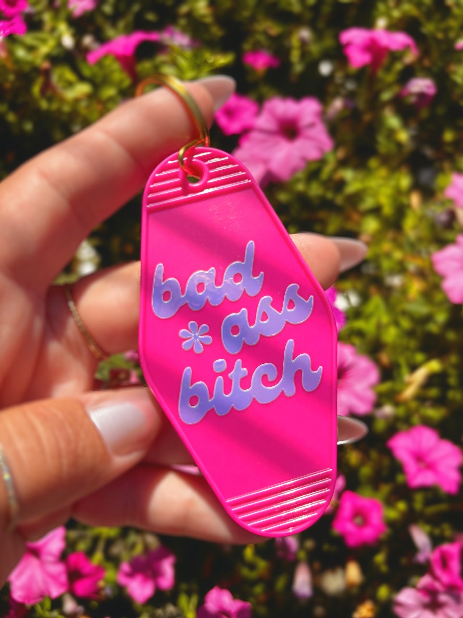 A hand holds a bright pink flip-flop-shaped keychain outdoors in front of green foliage and pink flowers. The keychain has the phrase &
