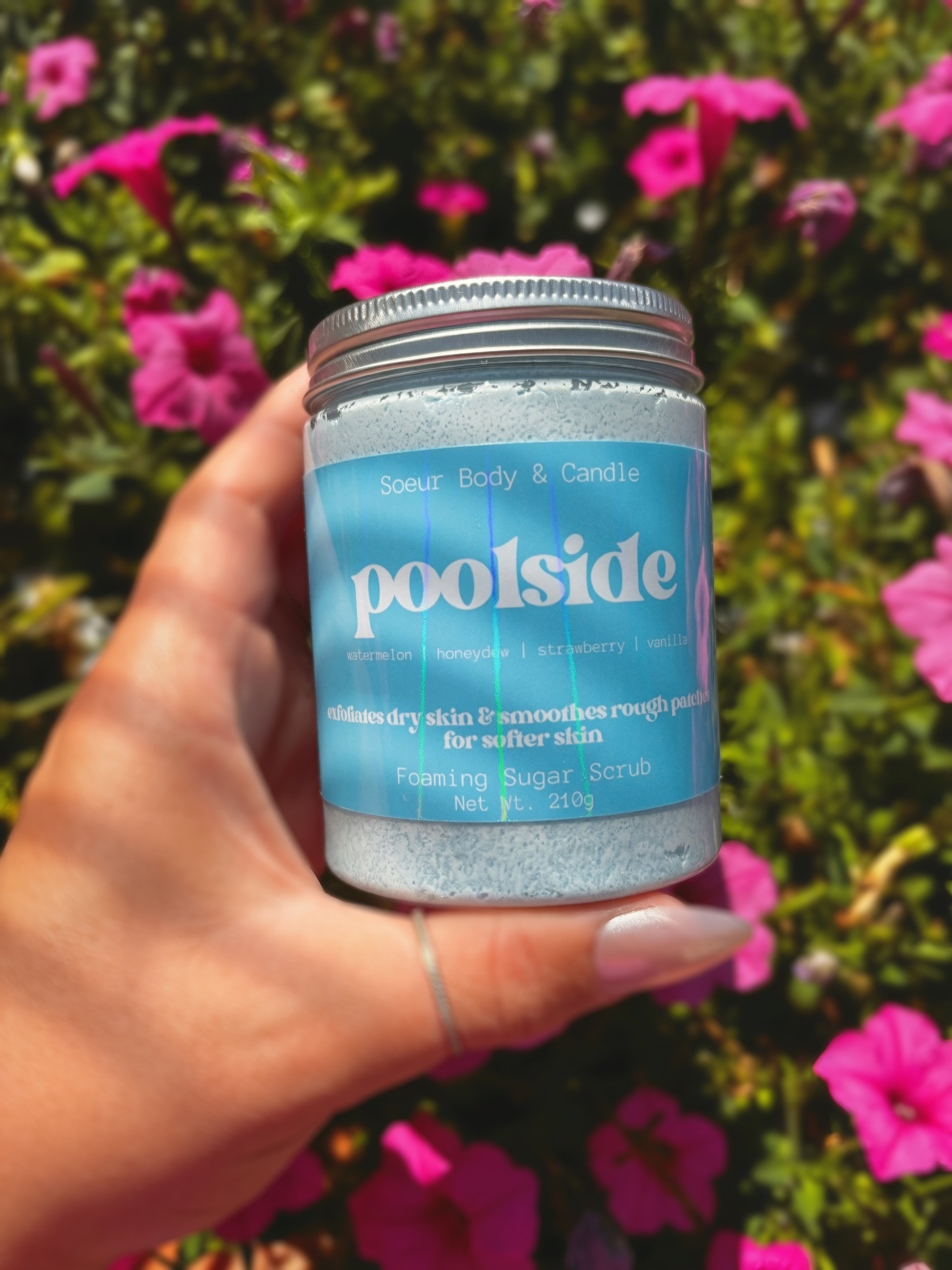 A hand holds a jar of Poolside Foaming Sugar Scrub against a background of green foliage and pink flowers. The jar has a light blue label with white text and a silver metal lid.
