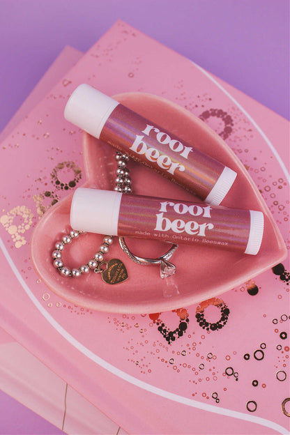 Two Root Beer flavoured lip balms displayed in a jewelry dish with a ring and bracelet, sitting on top of books.