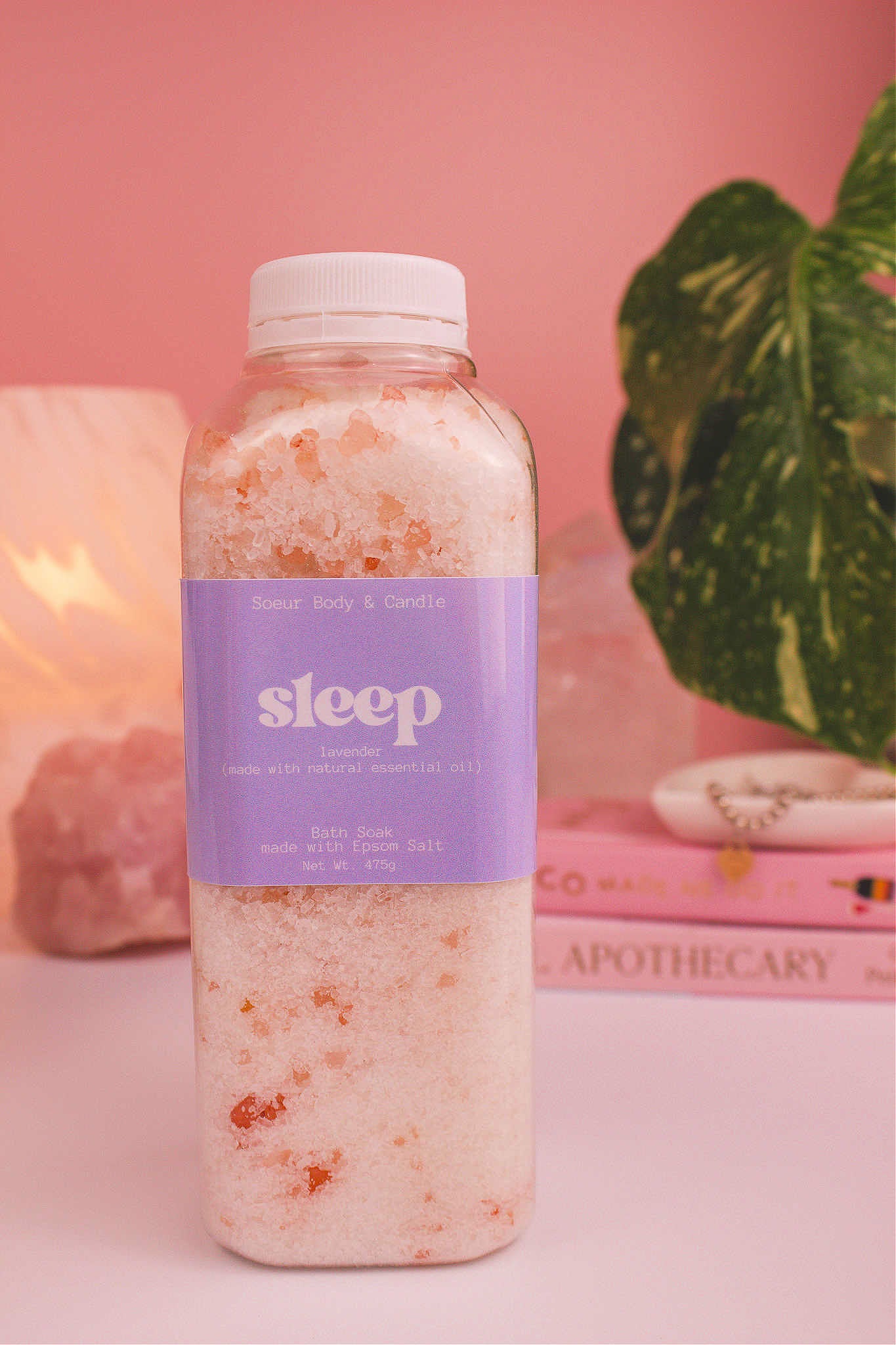 Sleep bath soak with a stack of books, a jewelry dish, lamp, plants, and crystals in the background; made with lavender essential oil.