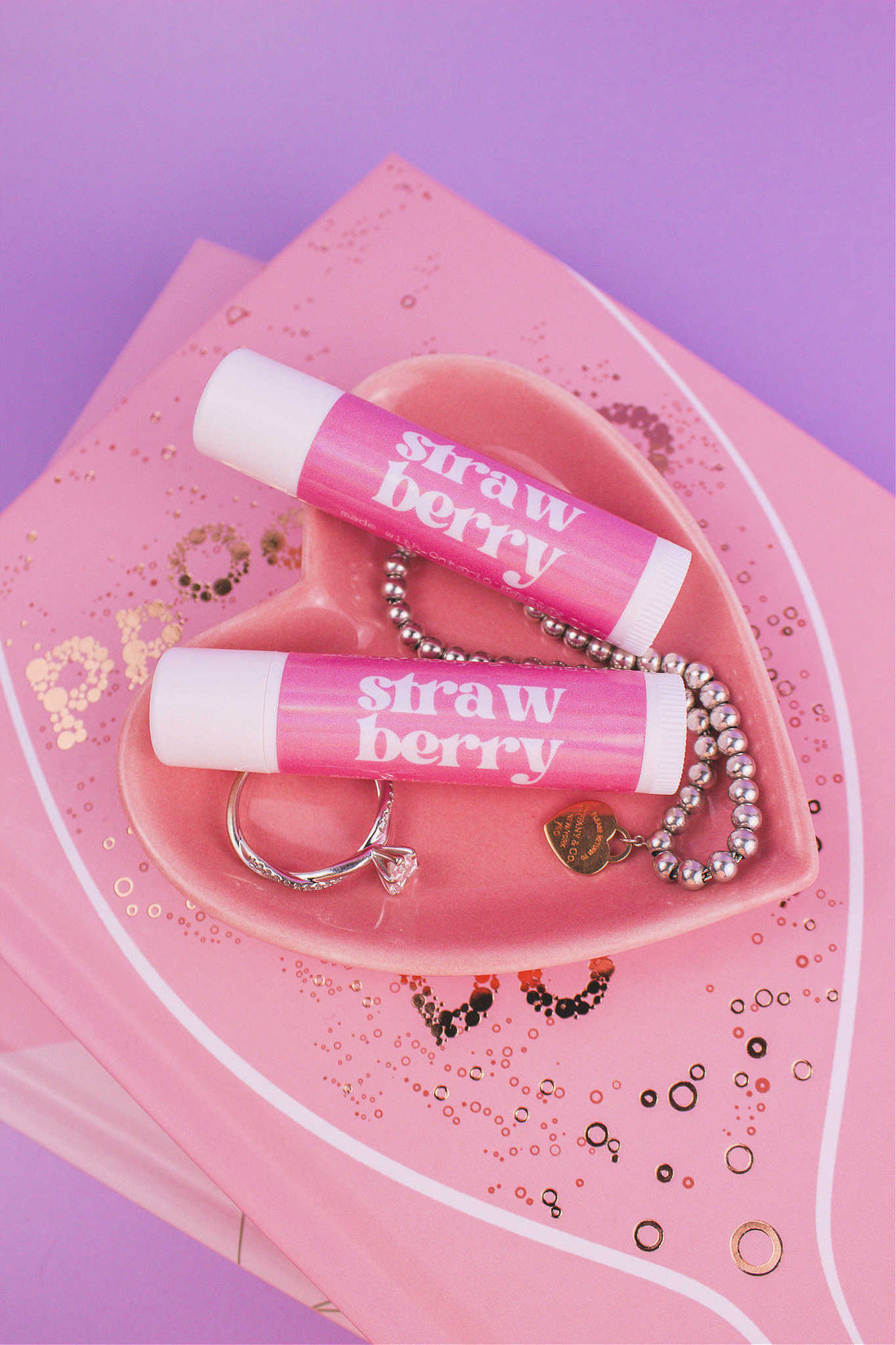 Two Strawberry flavoured lip balms displayed in a jewelry dish with a ring and bracelet, sitting on top of books.