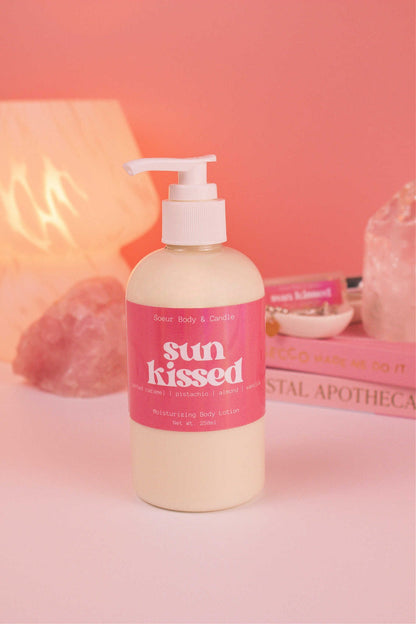 Sun Kissed body lotion with a stack of books, a jewelry dish, lamp, and crystals in the background; fragrance notes: salted caramel, pistachio, almond, and vanilla.