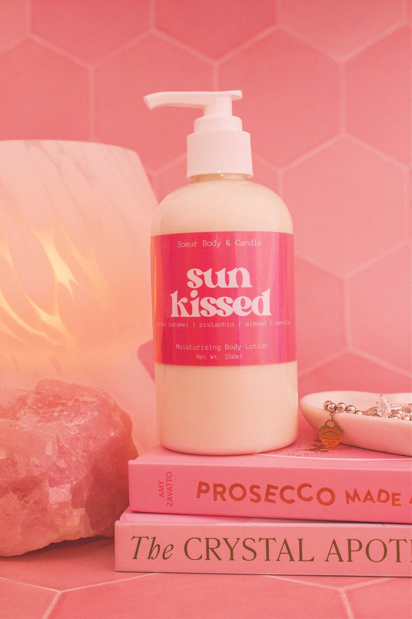 Close-up of the Sun Kissed body lotion on a stack of books with a jewelry dish, lamp, and crystal in the background; fragrance notes: salted caramel, pistachio, almond, and vanilla.