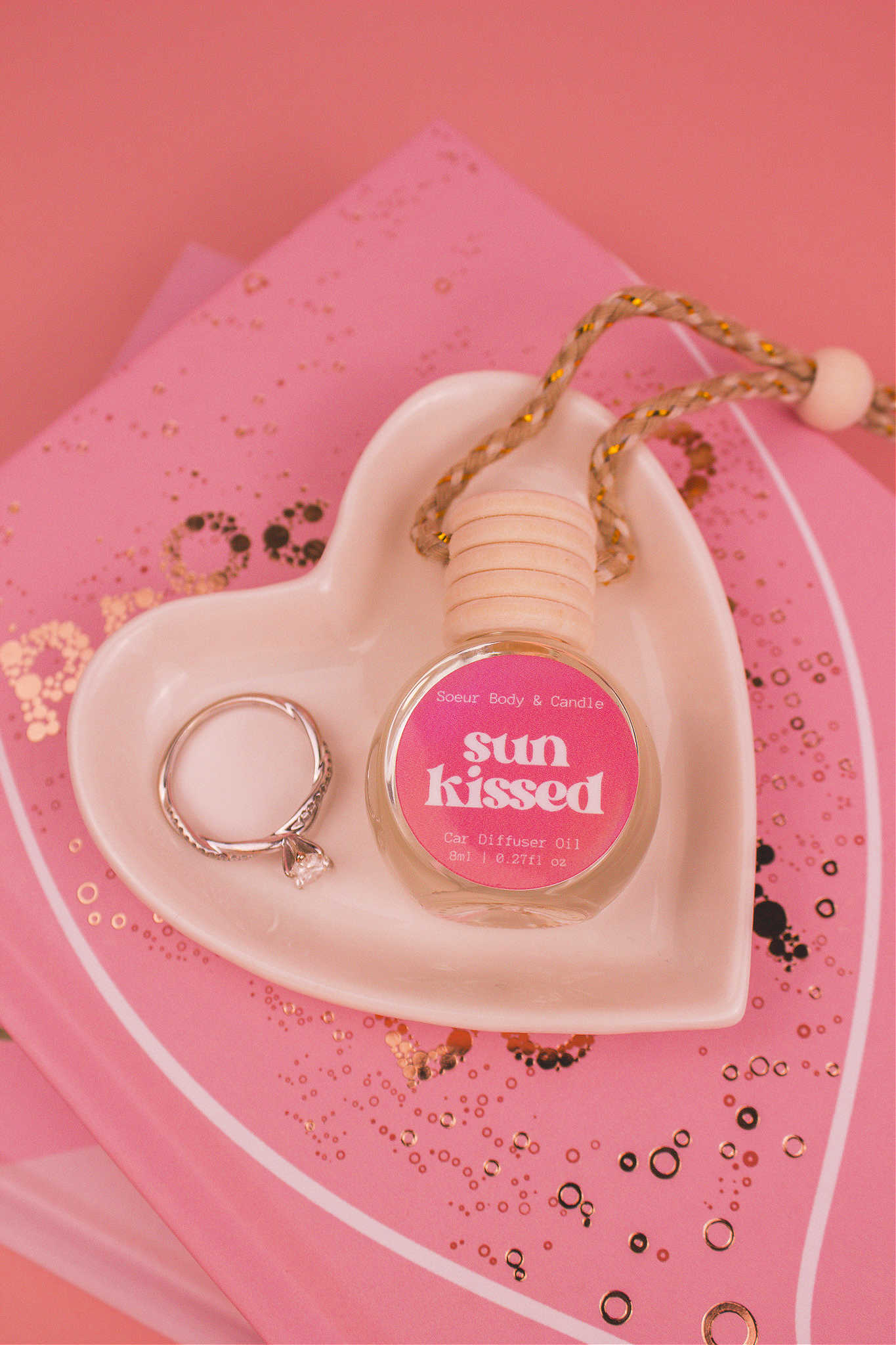 Sun Kissed car diffuser in a jewelry dish with a ring beside it, sitting on top of books, featuring notes of salted caramel, pistachio, almond, and vanilla.