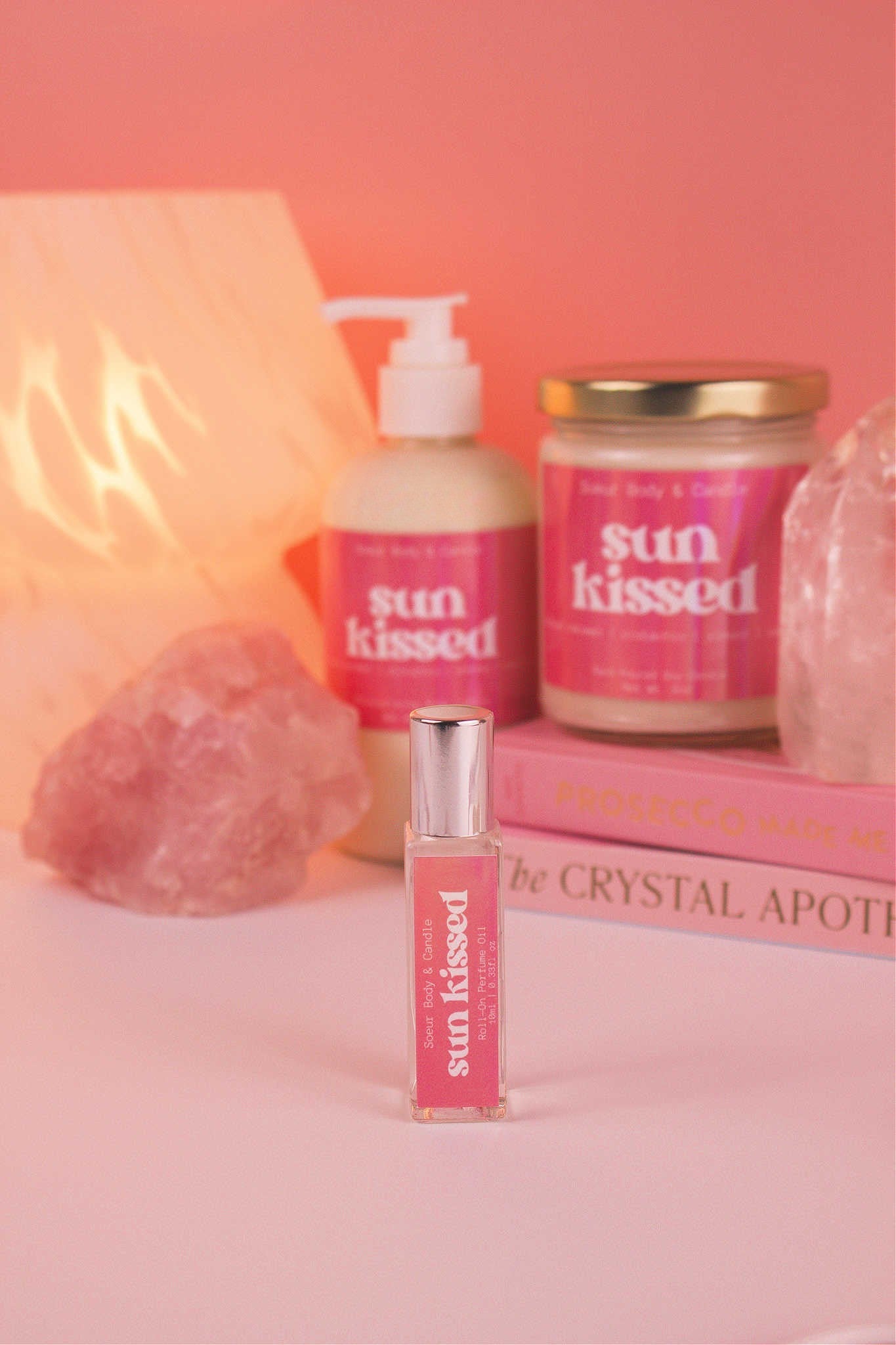 Focused shot of Sun Kissed roll-on perfume in the foreground with its matching lotion and candle, along with books, a lamp, and crystals softly blurred in the background. Featuring notes of salted caramel, pistachio, almond, and vanilla.