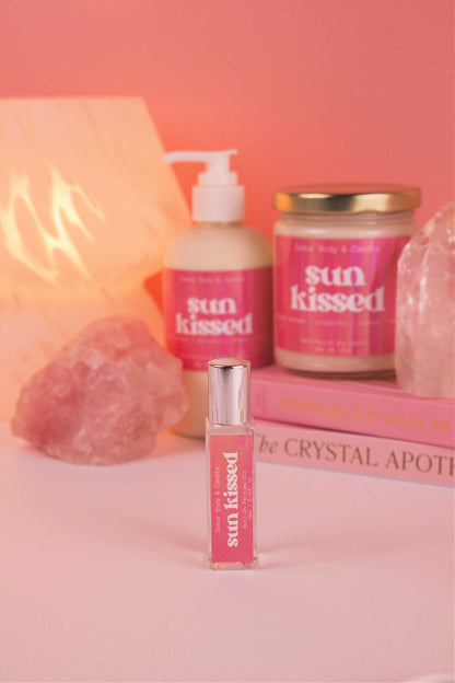 Focused shot of Sun Kissed roll-on perfume in the foreground with its matching lotion and candle, along with books, a lamp, and crystals softly blurred in the background. Featuring notes of salted caramel, pistachio, almond, and vanilla.