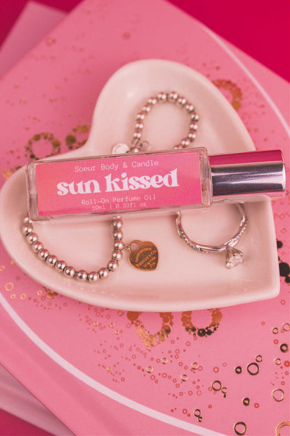 Close-up of Sun Kissed roll-on perfume bottle displayed in a jewelry dish beside a ring and bracelet, on top of books. Featuring notes of salted caramel, pistachio, almond, and vanilla.