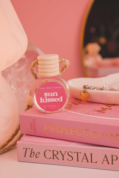 Sun Kissed diffuser sitting on top of a stack of books with a jewelry dish in the background, set against a lamp, mirror, and crystals; fragrance notes: salted caramel, pistachio, almond, and vanilla.