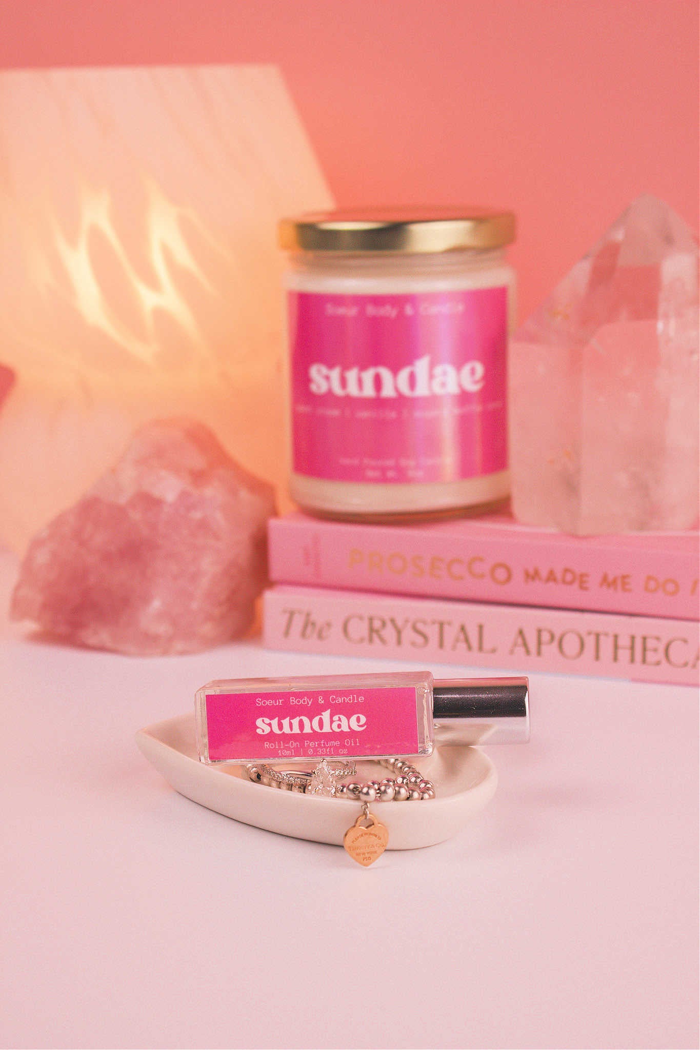 Sundae roll-on perfume elegantly set in a jewelry dish with jewelry, and coordinating candle, along with books, a lamp, and crystals in the background. Featuring notes of sweet cream, vanilla, and sugary waffle cone.