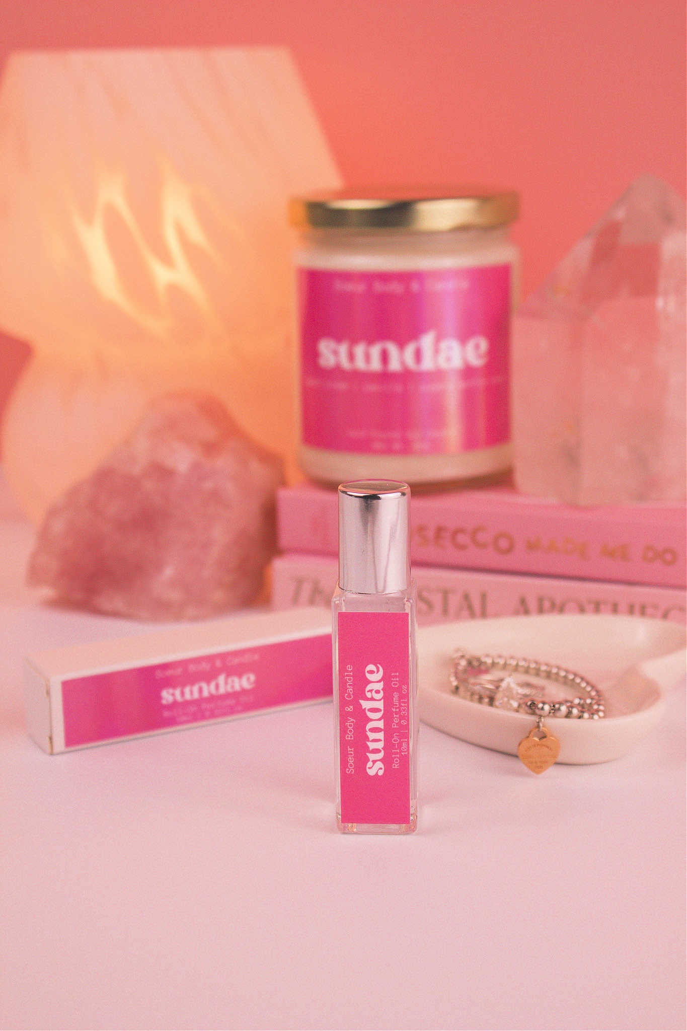 Focused shot of Sundae roll-on perfume in the foreground with its matching candle, along with a jewelry dish, books, a lamp, and crystals softly blurred in the background. Featuring notes of sweet cream, vanilla, and sugary waffle cone.