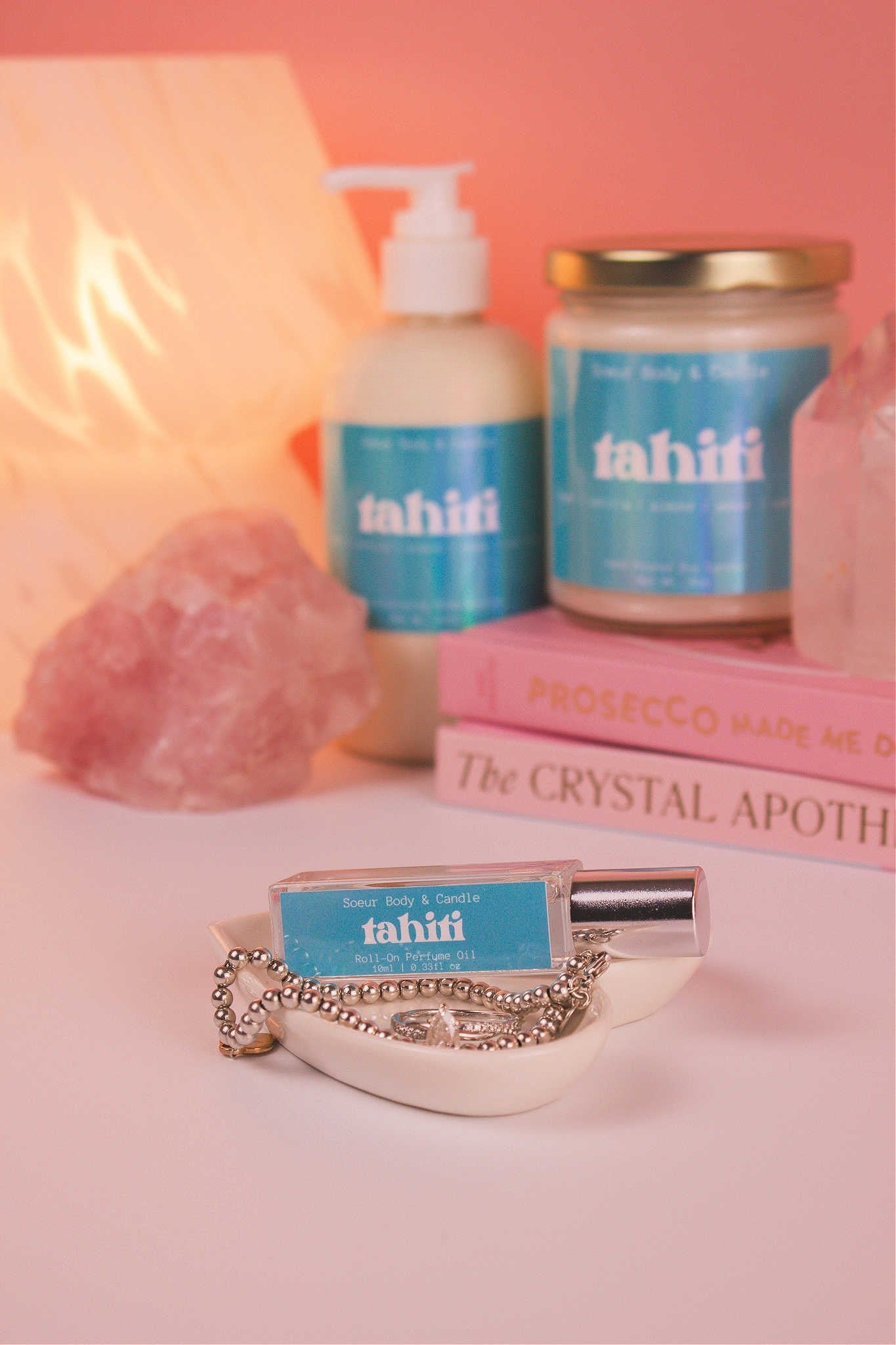 Tahiti roll-on perfume sitting elegantly in a jewelry dish with jewelry, and coordinating lotion and candle, along with books, a lamp, and crystals in the background. Featuring a blend of sugar, vanilla, almond, amber, and oak moss notes.