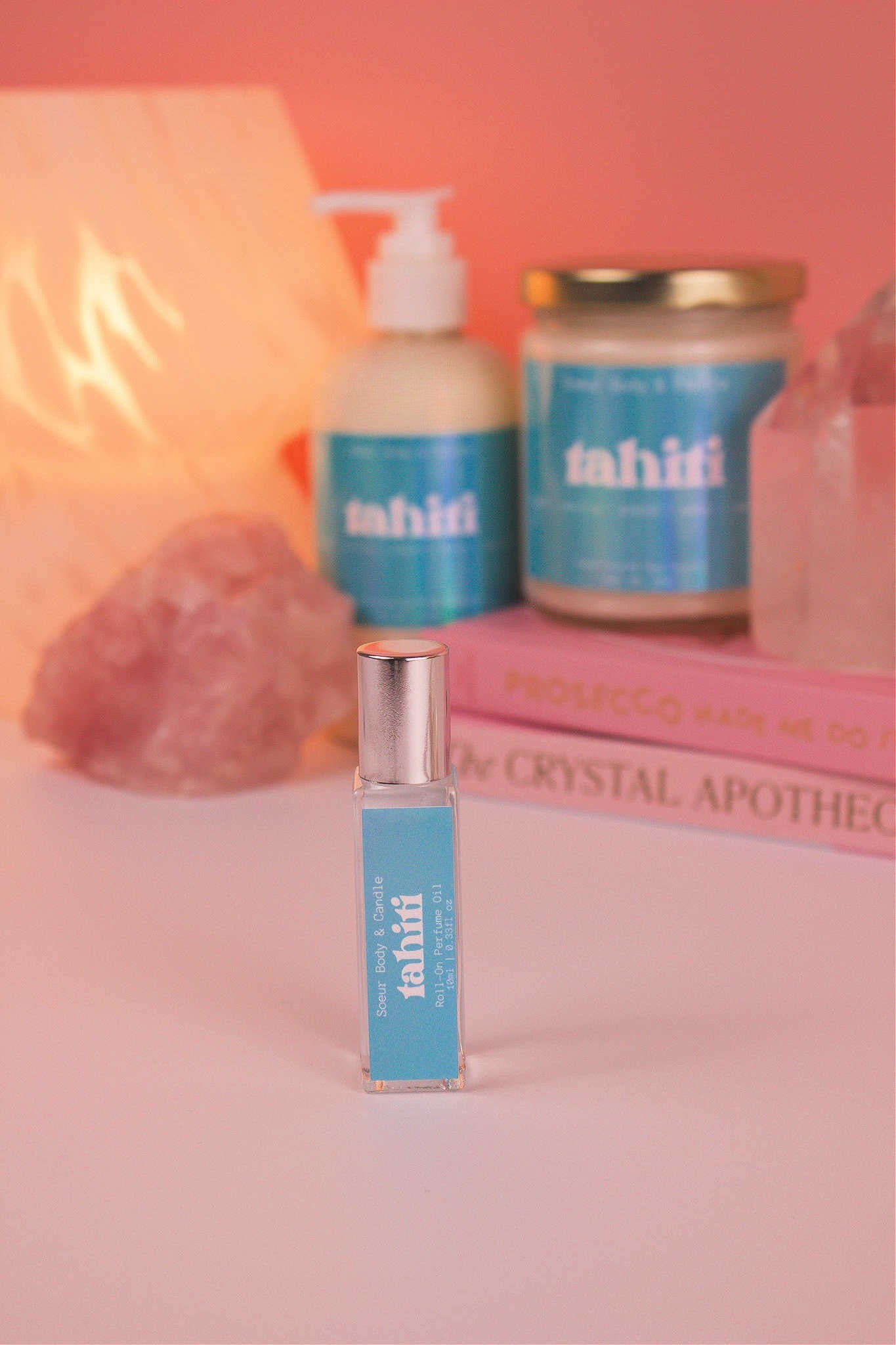 Focused shot of Tahiti roll-on perfume in the foreground with its matching lotion and candle, along with books, a lamp, and crystals softly blurred in the background. Featuring a sweet blend of sugar, vanilla, almond, amber, and oak moss notes.