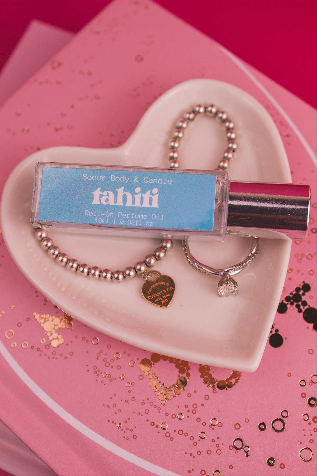 Tahiti roll-on perfume bottle displayed in a jewelry dish beside a ring and bracelet. Featuring exotic notes of sugar, vanilla, almond, amber, and oak moss.