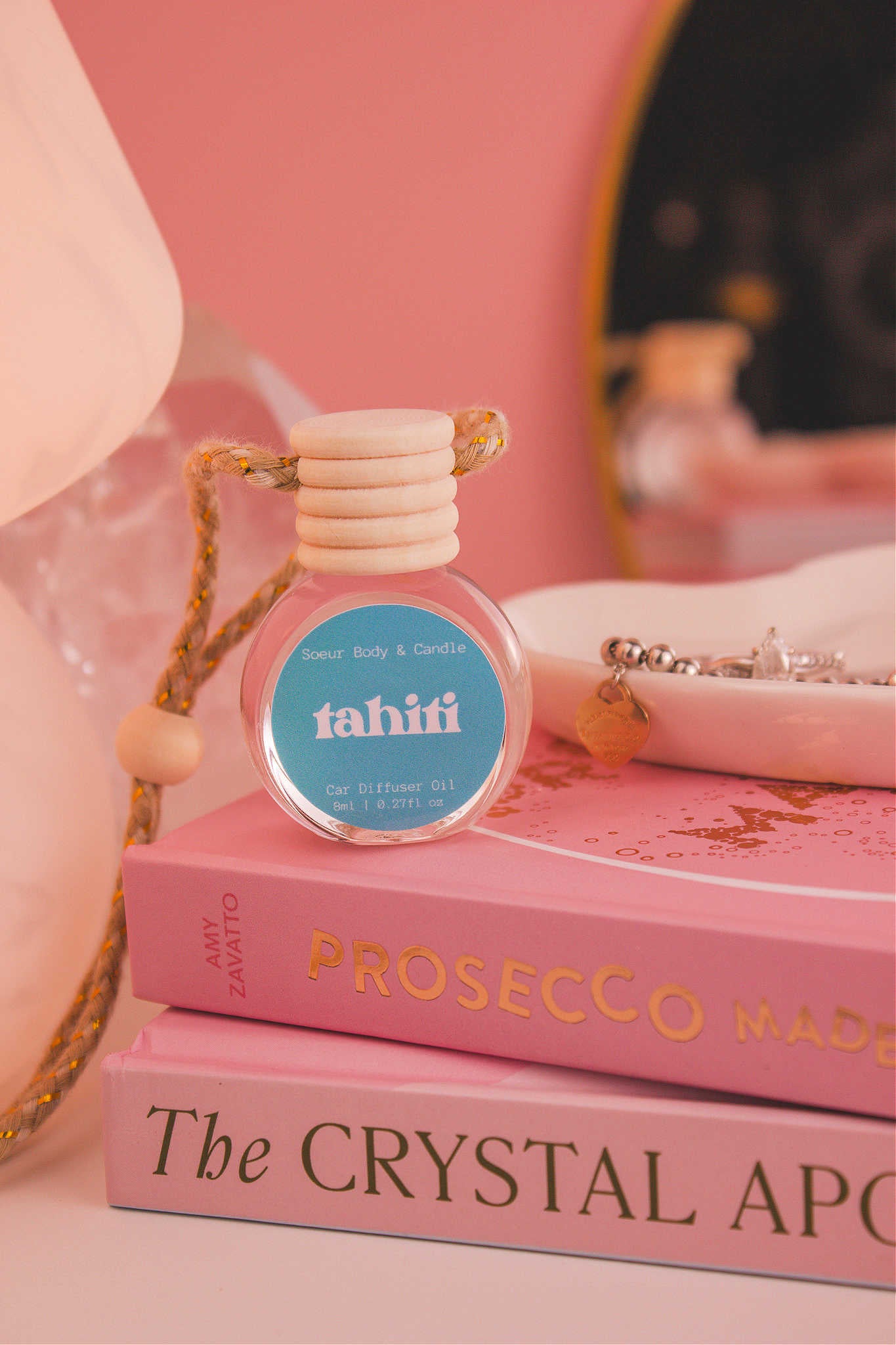 Tahiti diffuser sitting on top of a stack of books with a jewelry dish in the background, set against a lamp, mirror, and crystals; fragrance notes: sugar, vanilla, almond, amber, and oak moss.