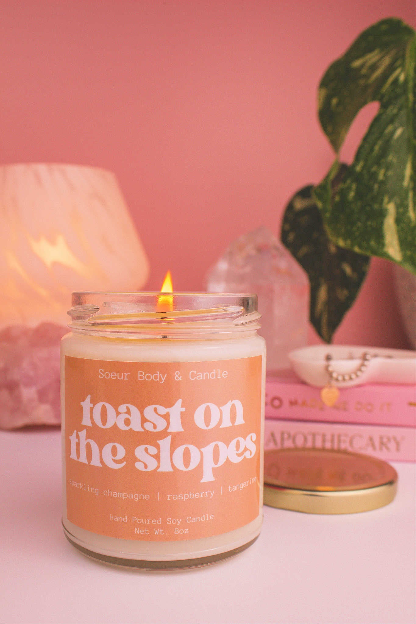 Close-up of Toast on the Slopes candle lit, with a stack of books and a jewelry dish in the background, set against a lamp, plant, and crystals; fragrance notes: sparkling champagne, raspberry, and tangerine.
