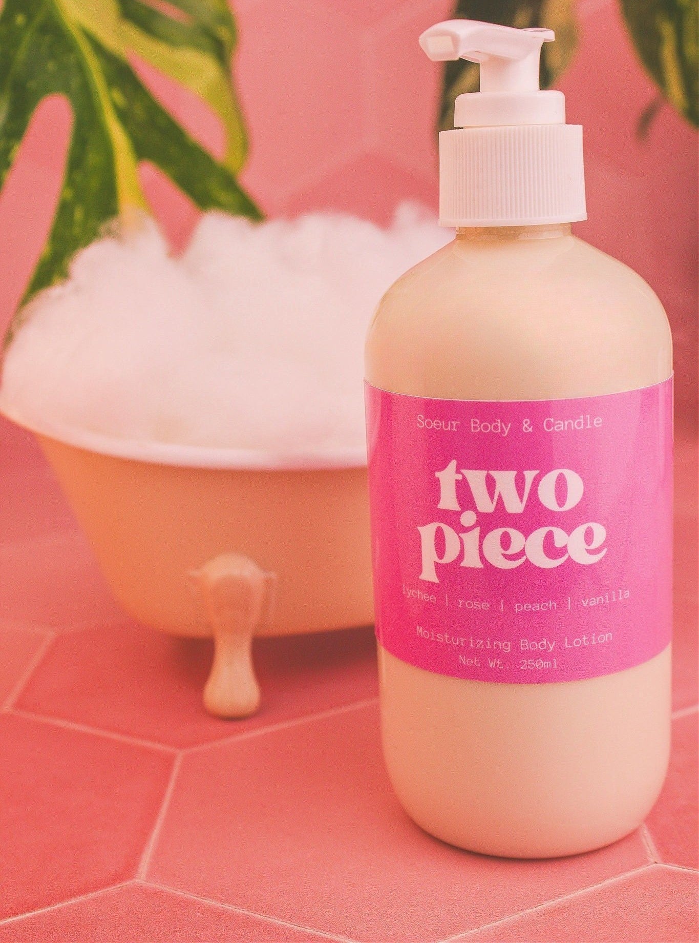 Two Piece body lotion in front of a small tub with bubbles and plants in the background, evoking a relaxing vibe with lychee, rose, peach, and vanilla notes.