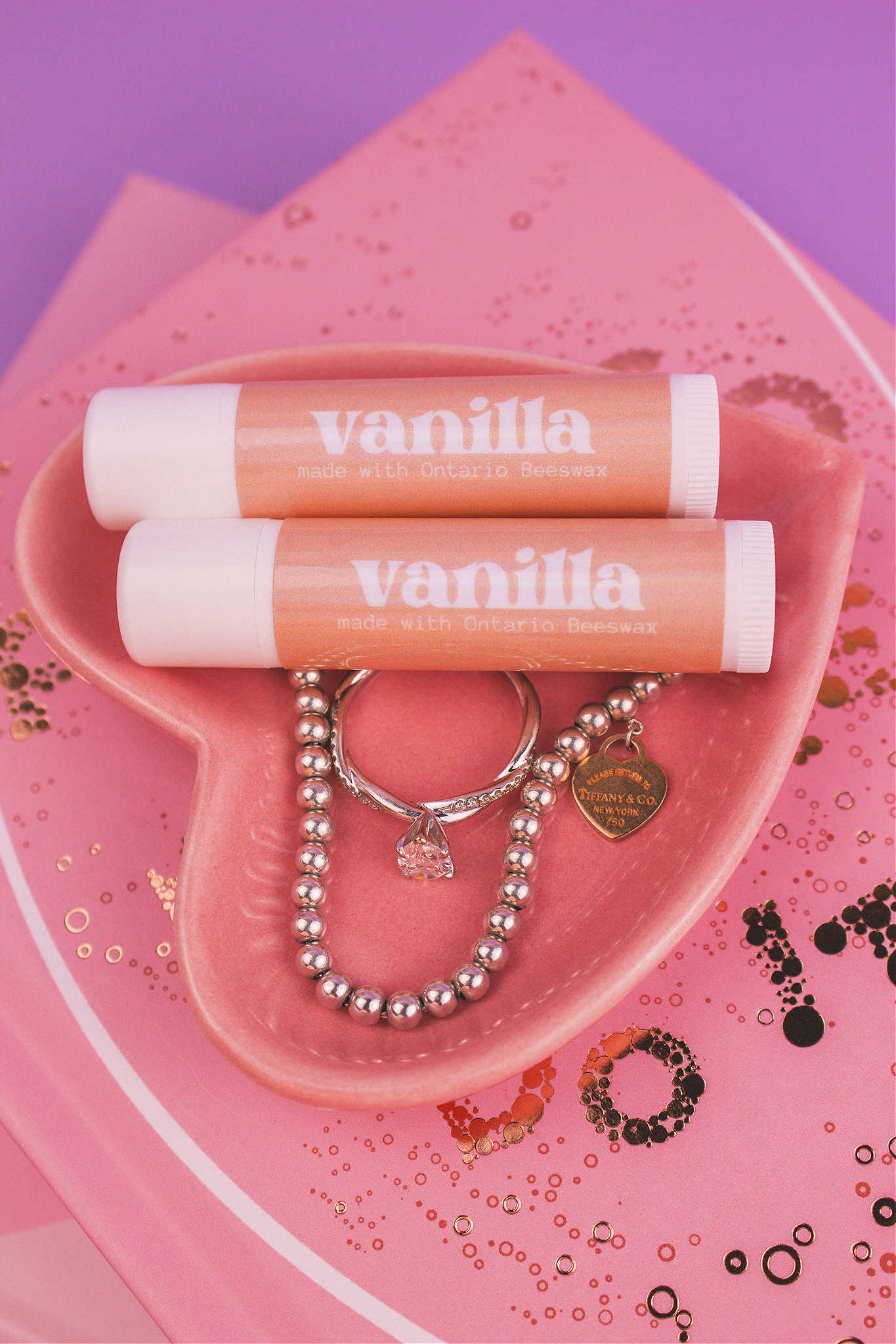 Two Vanilla flavoured lip balms displayed in a jewelry dish with a ring and bracelet, sitting on top of books.