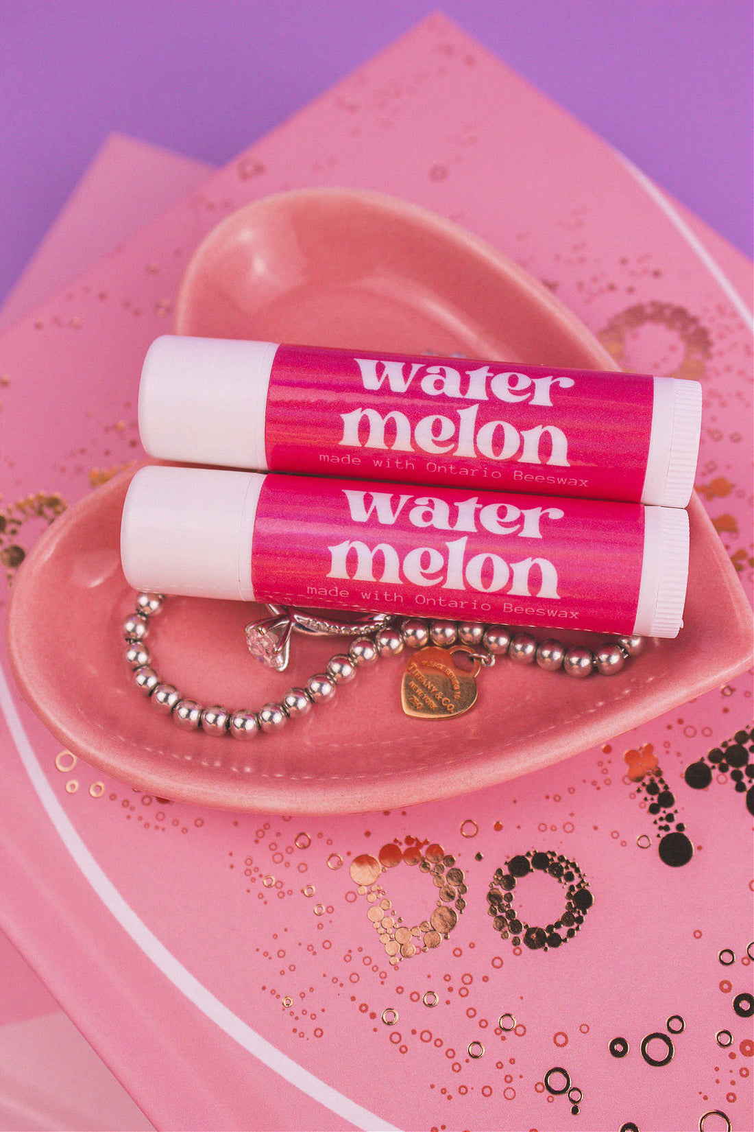 Two Watermelon flavoured lip balms displayed in a jewelry dish with a ring and bracelet, sitting on top of books.