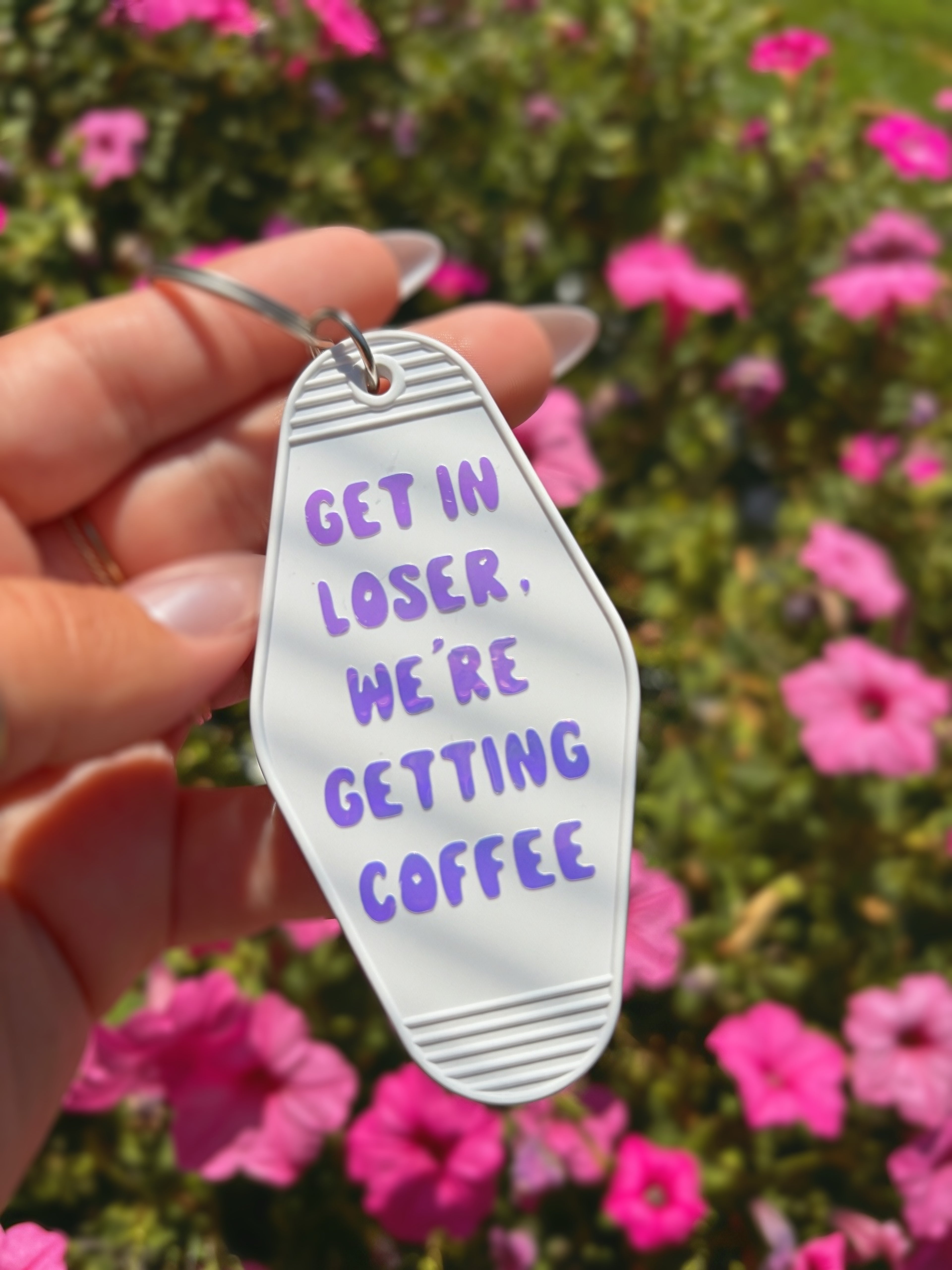 A hand holds a white shoe-shaped keychain with white holographic text that reads &
